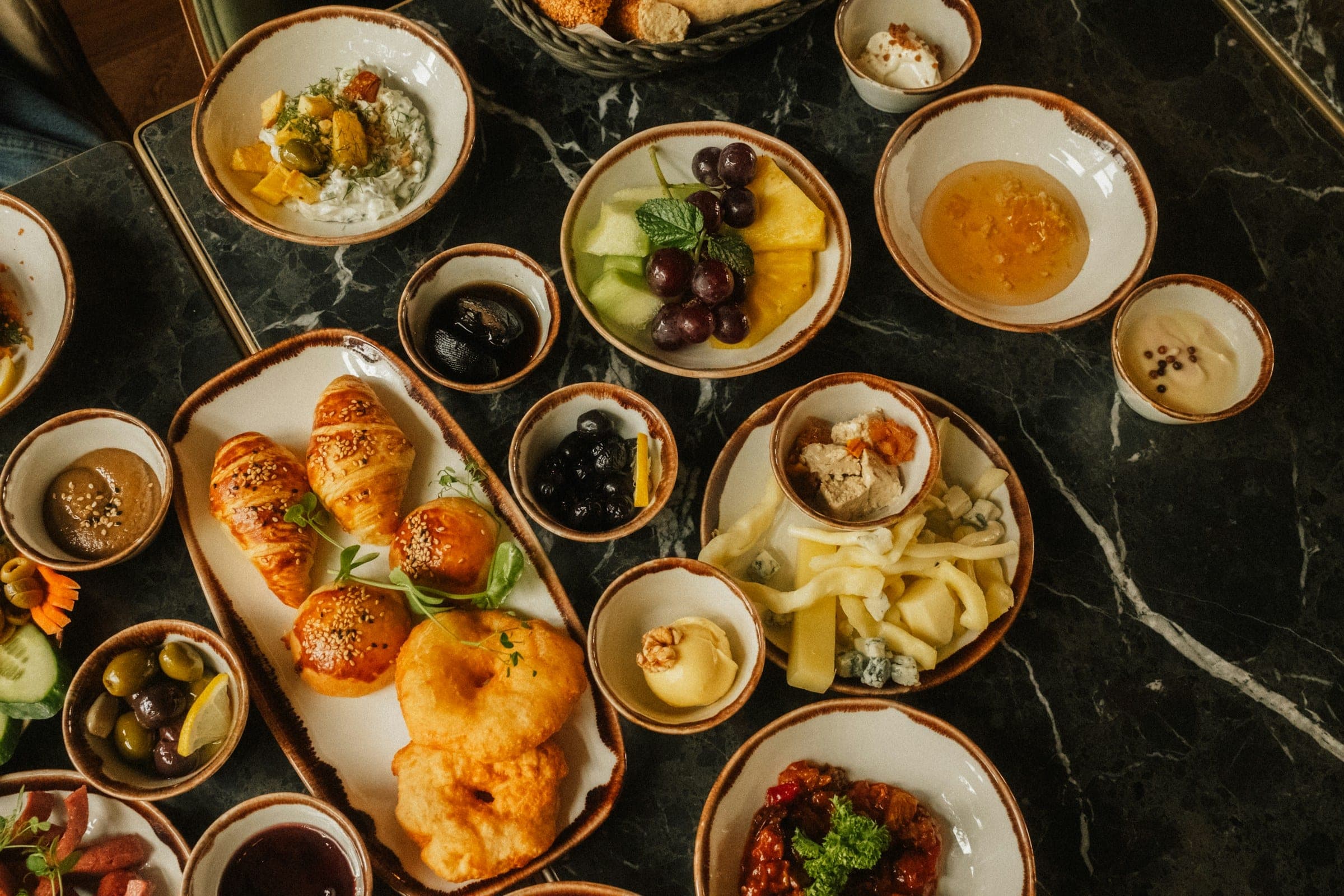 Overhead view of a breakfast spread of sesame pastries, fruit, cheeses, olives and preserves in small bowls on a dark marble table