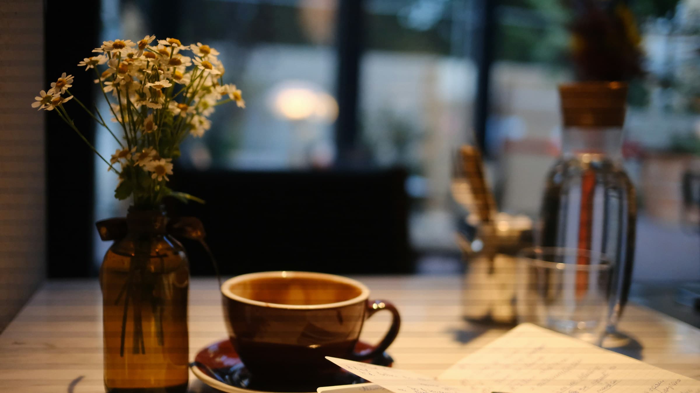 A cup of coffee on a saucer beside a small vase of daisies on a table in soft dusk light