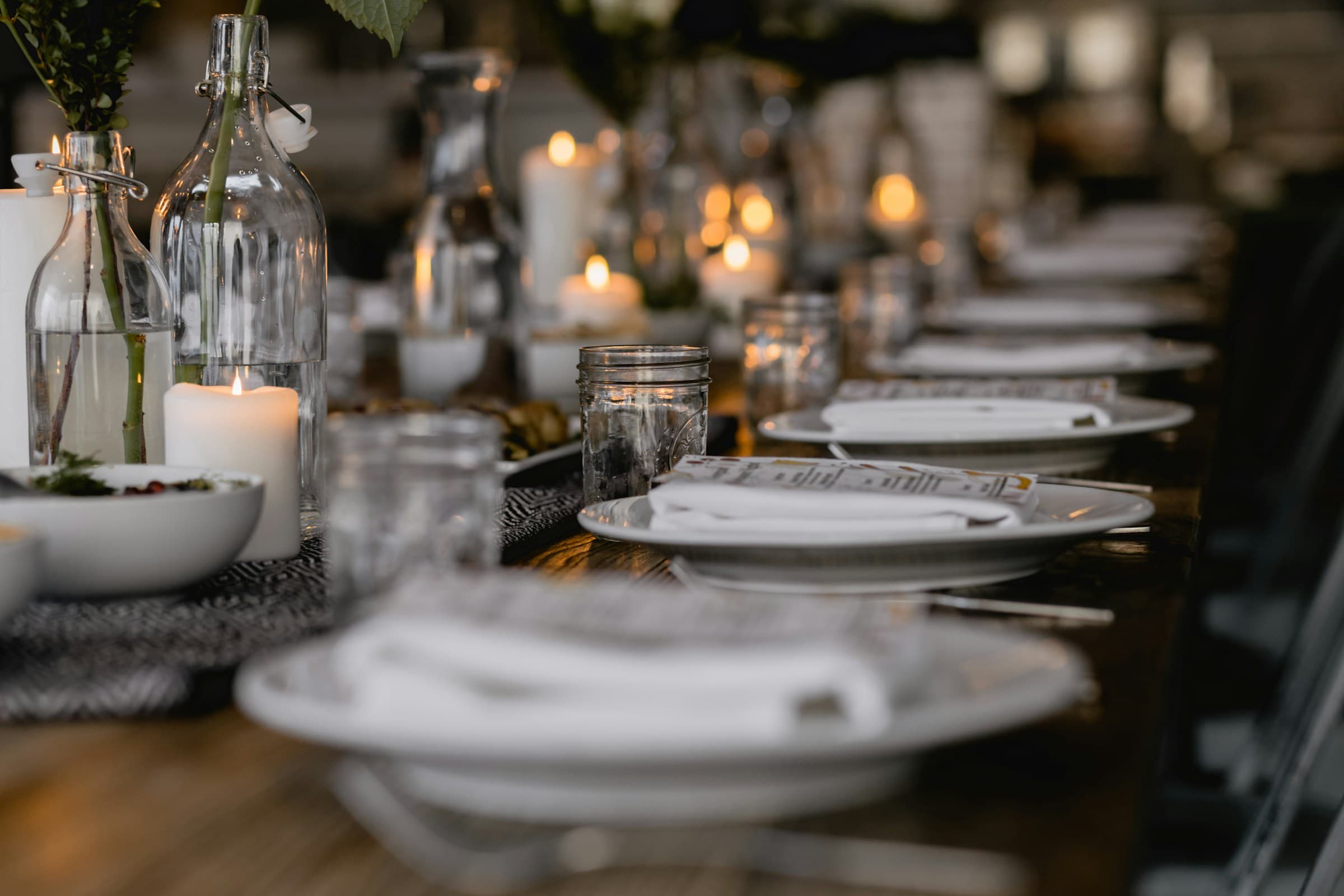 A long dinner table set with white plates, folded napkins and glass bottles, lit by rows of candles in warm evening light